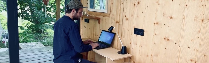 Person arbeitet an einem Laptop an einem kleinen Holzschreibtisch in einem minimalistischen Tiny-House-Interieur; durch die offene Glastür ist eine grüne Waldlandschaft zu sehen.