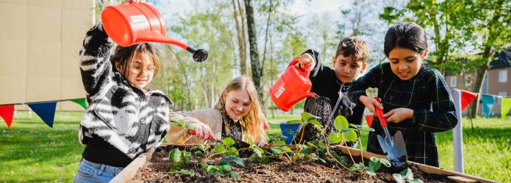 Vier Kinder gärtnern gemeinsam an einem Hochbeet im Freien: Sie gießen junge Pflanzen mit roten Gießkannen und arbeiten mit kleinen Gartengeräten, umgeben von Grünflächen und Bäumen.