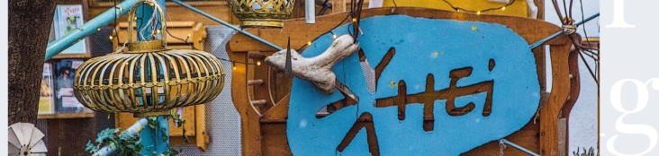 Ein brauner ehemaliger Lampenschirm aus Bambus hängt vor einer Art Holztor, worauf ein blaues Schild mit der Aufschrift HEI zu sehen ist.