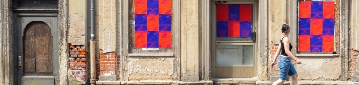 Eine Frau läuft sommerlich gekleidet an einem sanierungsbedürftigem hellbraunem Haus vorbei. Die drei ehemaligen Fenster und Türen sind mit Karomusterplakaten in orange/blau beklebt.