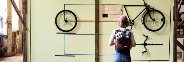 Person mit Rucksack und Fahrradhelm steht in einer Industriehalle vor einer großformatigen Wandinstallation, an der Fahrradteile wie Rahmen, Räder und Lenker grafisch angeordnet sind, ähnlich einer technischen Zeichnung.