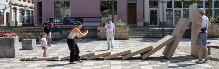 Menschen auf einem öffentlichen Platz interagieren mit einer bodennah aufgebauten Kette aus großen, rechteckigen Kartonelementen, die wie fallende Dominosteine angeordnet sind; im Hintergrund stehen bunte Stadthäuser und Bäume.