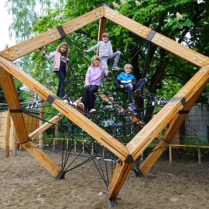 Drei Kinder, die auf einer großen, begehbaren Holzwürfel-Kletterstruktur aus Balken stehen und spielen, auf sandigem Untergrund.
