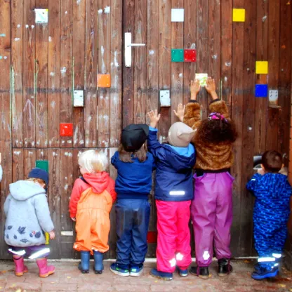 6 Kinder stehen vor einem Holz-Gartentor einer Kita und verschönern es mit bunten Etiketten.