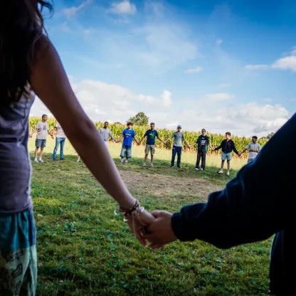 Zwei Personen halten sich im Vordergrund auf einer Wiese an den Händen. Im Hintergrund bildet eine Gruppe junger Menschen einen Kreis, ebenfalls Hand in Hand, vor einem Maisfeld und blauem Himmel.