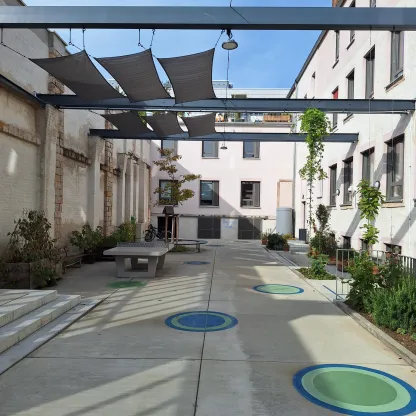 Zu sehen ist der neu entstandene Hinterhof des Integrativen Familienzentrums mit Tischtennisplatte, Verschattung und Begrünung. Auf dem Boden bunte gemalte Kreise in Blau und Grün.