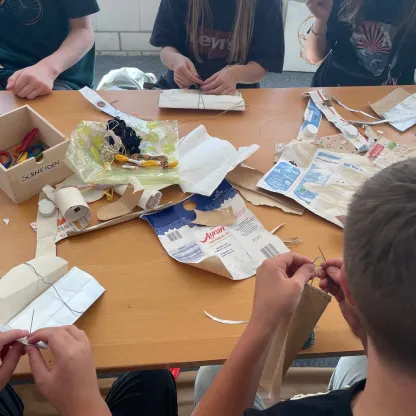 Hier ist ein Foto von einem Tisch zu sehen, an dem Kinder sitzen und aus Müll etwas Neues schaffen, sie nähen mit Nadel und Faden aus Milchverpackungen neue Produkte.
