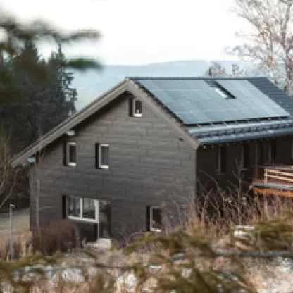 Bild des Bärenhaisla Jugendhauses: ein dunkelgraues Holzhaus mit Solarpanelen auf dem Dach, umgeben von Bäumen.
