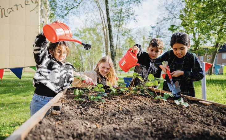 Vier Kinder gießen und pflegen gemeinsam Pflanzen in einem Hochbeet im Freien. Zwei von ihnen halten rote Gießkannen, ein Kind benutzt eine kleine Schaufel, während eine weiteres lächelnd die Erde harkt. Im Hintergrund sind Bäume und bunte Wimpelketten zu sehen.