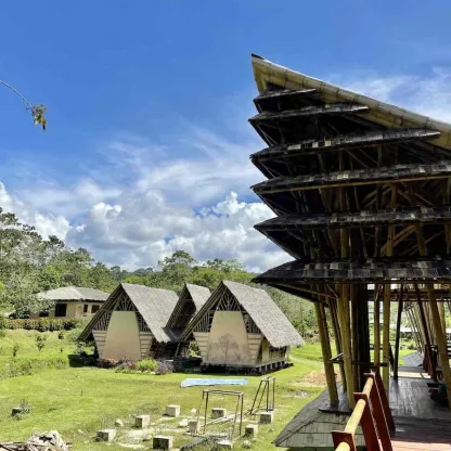 Nachhaltige Holzbauten in einer grünen Landschaft: Mehrere kleine Häuser mit steilen, traditionellen Dächern stehen auf einer Wiese, im Vordergrund ein offener Pavillon aus Holz unter blauem Himmel.