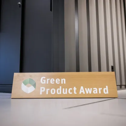 Holzschild mit dem Schriftzug „Green Product Award" und dem zugehörigen Logo – einem geometrischen, blattförmigen Symbol in Grün- und Weißtönen – lehnt auf einem hellen Betonboden vor einer dunklen Schrankwand mit vertikalen Lamellen im Hintergrund.