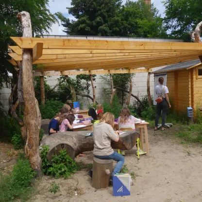 Mehrere Schulkinder sitzend, die in einem outdoor-Klassenzimmer an Möbeln aus Holz arbeiten, eine Lehrerin steht an einer Tafel und notiert etwas darauf, die Unterrichtsfläche ist mit einem Holzdach überdacht, welches bis zu einer Hütte aus Holz reicht.