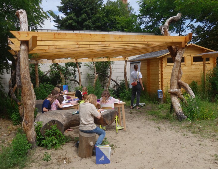 Mehrere Schulkinder sitzend, die in einem outdoor-Klassenzimmer an Möbeln aus Holz arbeiten, eine Lehrerin steht an einer Tafel und notiert etwas darauf, die Unterrichtsfläche ist mit einem Holzdach überdacht, welches bis zu einer Hütte aus Holz reicht.