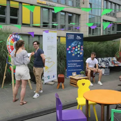 Infostand auf einer öffentlichen Veranstaltung: Mehrere Menschen kommen miteinander ins Gespräch, neben einem Glücksrad und Informationsbannern zu Nachhaltigkeit und Digitalisierung; bunte Wimpelketten hängen über Sitzgelegenheiten und einem kleinen Pavillon im urbanen Außenraum.