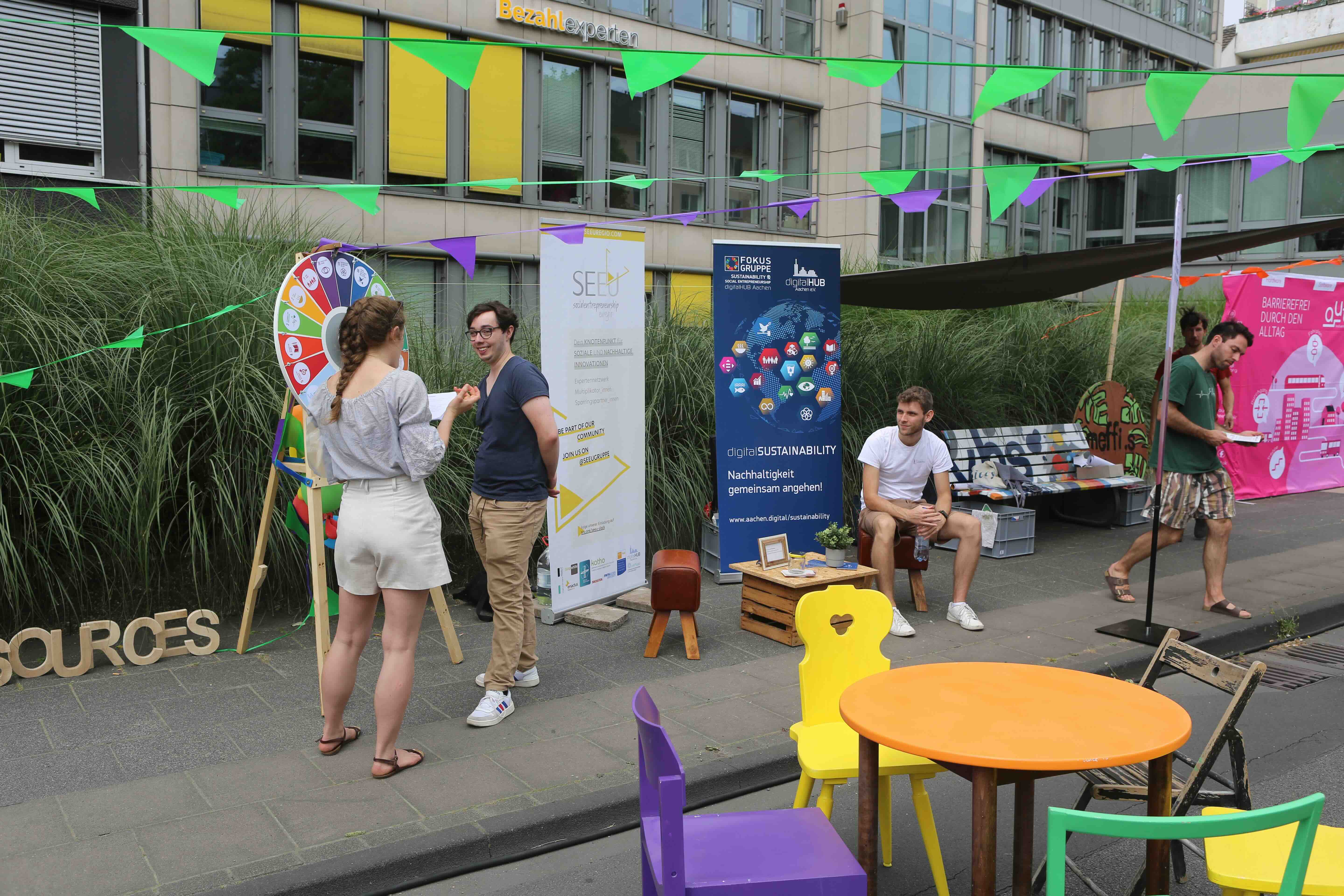Infostand auf einer öffentlichen Veranstaltung: Mehrere Menschen kommen miteinander ins Gespräch, neben einem Glücksrad und Informationsbannern zu Nachhaltigkeit und Digitalisierung; bunte Wimpelketten hängen über Sitzgelegenheiten und einem kleinen Pavillon im urbanen Außenraum.