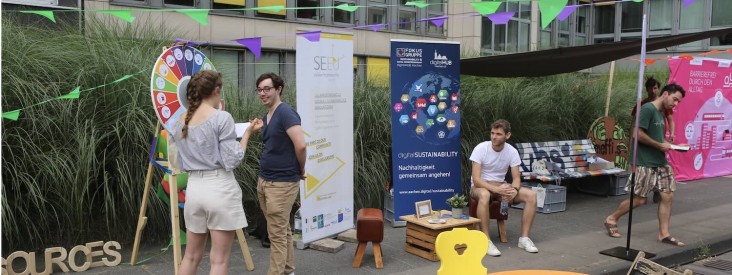 Infostand auf einer öffentlichen Veranstaltung: Mehrere Menschen kommen miteinander ins Gespräch, neben einem Glücksrad und Informationsbannern zu Nachhaltigkeit und Digitalisierung; bunte Wimpelketten hängen über Sitzgelegenheiten und einem kleinen Pavillon im urbanen Außenraum.