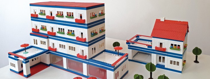 Modell eines mehrstöckigen Wohn- und Geschäftskomplexes aus einem Architekturbaukasten: Weiße Gebäude mit roten Dächern, blauen Fassadenstreifen, Balkonen und kleinen Bäumen sind zu einer blockartigen Anlage arrangiert.