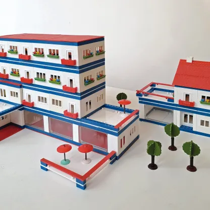 „Modell eines mehrstöckigen Wohn- und Geschäftskomplexes aus einem Architekturbaukasten: Weiße Gebäude mit roten Dächern, blauen Fassadenstreifen, Balkonen und kleinen Bäumen sind zu einer blockartigen Anlage arrangiert.