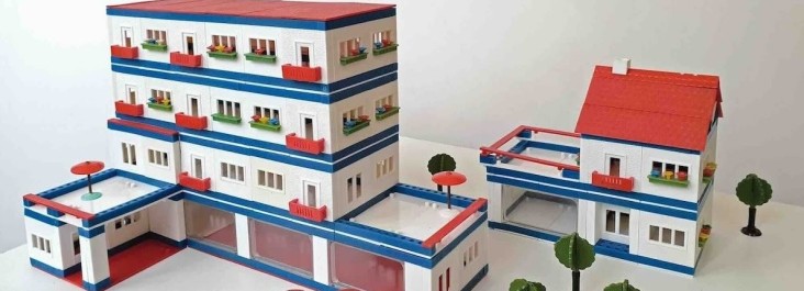 „Modell eines mehrstöckigen Wohn- und Geschäftskomplexes aus einem Architekturbaukasten: Weiße Gebäude mit roten Dächern, blauen Fassadenstreifen, Balkonen und kleinen Bäumen sind zu einer blockartigen Anlage arrangiert.