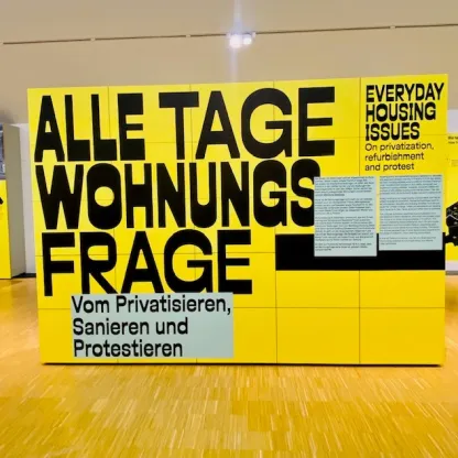 Zu sehen ist ein Ausstellungsraum mit aufgestellter Plakatwand, dort steht schwarz auf gelb: „Alle Tage Wohnungsfrage – Vom Privatisieren, Sanieren und Protextieren“ sowie der Titel der Ausstellung auf Englisch: „Everyday Housing Issues. On privatisation, refurbishment and protest.“