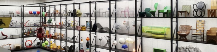 Auf dem Bild sind in einem schwarzen Regal vor einer weißen Wand verschiedene Ausstellungsexponate, z.B. verschiedene Sitzmöbel abgebildet.