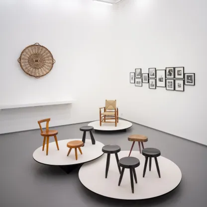 Museumsansicht mit mehreren Holzstühlen und -hockern auf runden, erhöhten Präsentationsplattformen sowie Kunstwerken und einem Flechtwerk an den Wänden.
