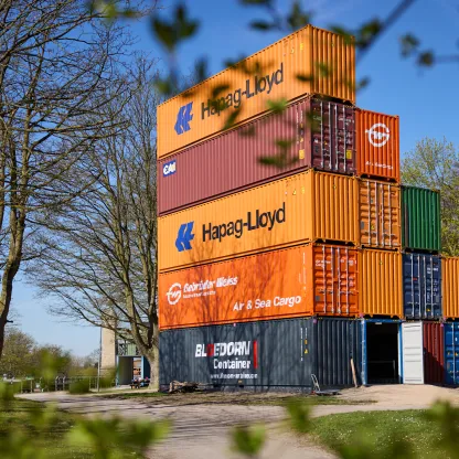 Foto von meheren treppenartig gestapelten bunten Schiffscontainern zwischen Bäumen und Büschen.