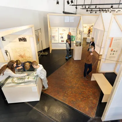 Mitte eine Nahaufnahme von zwei Personen, die sich über eine Vitrine beugen und ein Objekt genau betrachten, umgeben von Ausstellungstexten und Vitrinen.