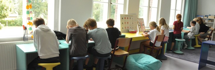 Kinder sitzen nebeneinander an langen Tischen in einem hellen Klassenraum und arbeiten konzentriert an Aufgaben; bunte Hocker, Stühle und Sitzkissen stehen vor großen Fenstern mit Tageslicht.