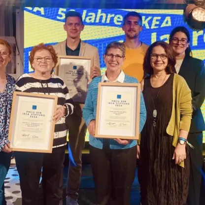Gruppenfoto der Preisträgerinnen und Preisträger des Preises der IKEA Stiftung 2024: Mehrere Personen stehen auf einer Bühne und halten gerahmte Urkunden der IKEA Stiftung, im Hintergrund eine beleuchtete Veranstaltungswand.