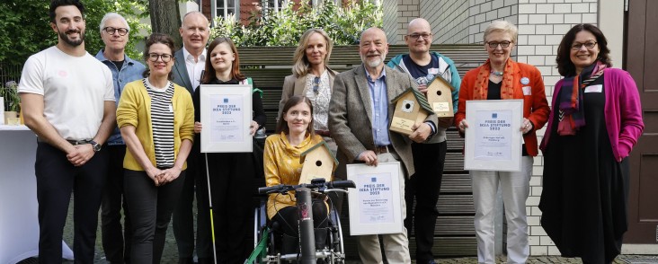 Gruppenfoto der Preisträgerinnen und Preisträger des Preises der IKEA Stiftung 2023: Mehrere Personen stehen vor einem Tor und halten gerahmte Urkunden der IKEA Stiftung, im Hintergrund ein Zaun und ein Haus.