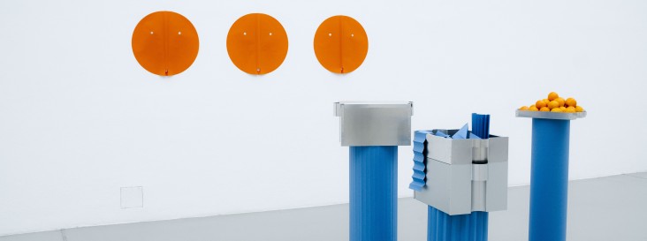Minimalistische Ausstellungsinstallation mit drei orangefarbenen, runden Wandobjekten und mehreren blauen Sockeln, auf denen metallische Behälter und eine Schale mit Orangen arrangiert sind, vor weißem Hintergrund.