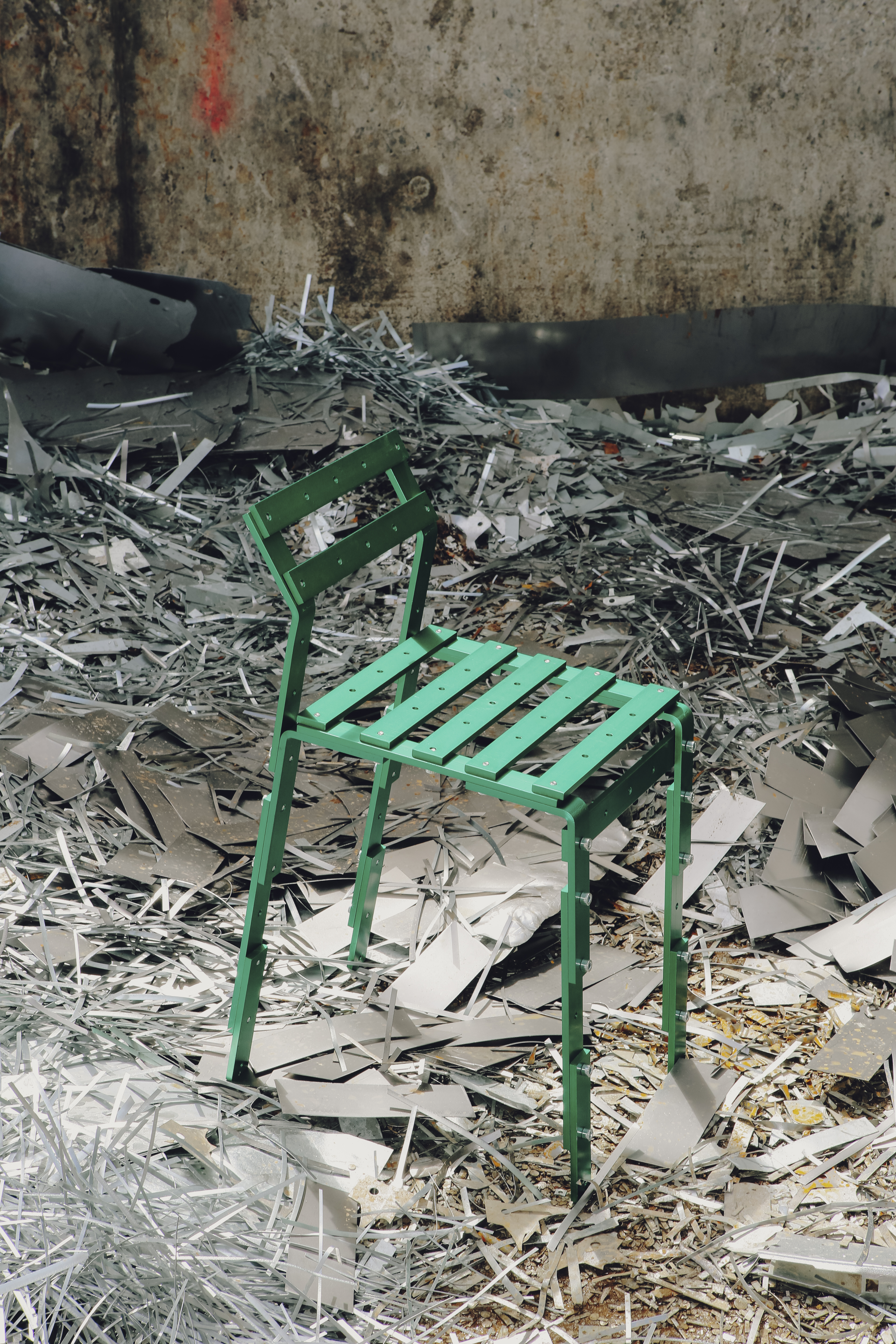Grüner Metallstuhl, designt von Luis Niederbuchner, mit Latten inmitten von Metallschrott – modernes Möbeldesign in roh-urbaner Industriekulisse.