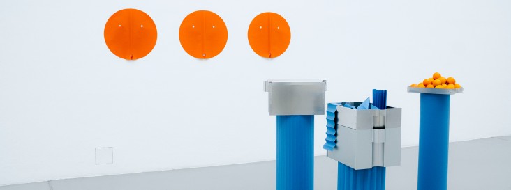 Minimalistische Ausstellungsinstallation mit drei orangefarbenen, runden Wandobjekten und mehreren blauen Sockeln, auf denen metallische Behälter und eine Schale mit Orangen arrangiert sind, vor weißem Hintergrund.