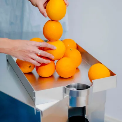 Hände stapeln Orangen auf einem minimalistischen Metallmöbel mit integrierten zylindrischen Öffnungen, modernes Produktdesign in Studio-Umgebung.