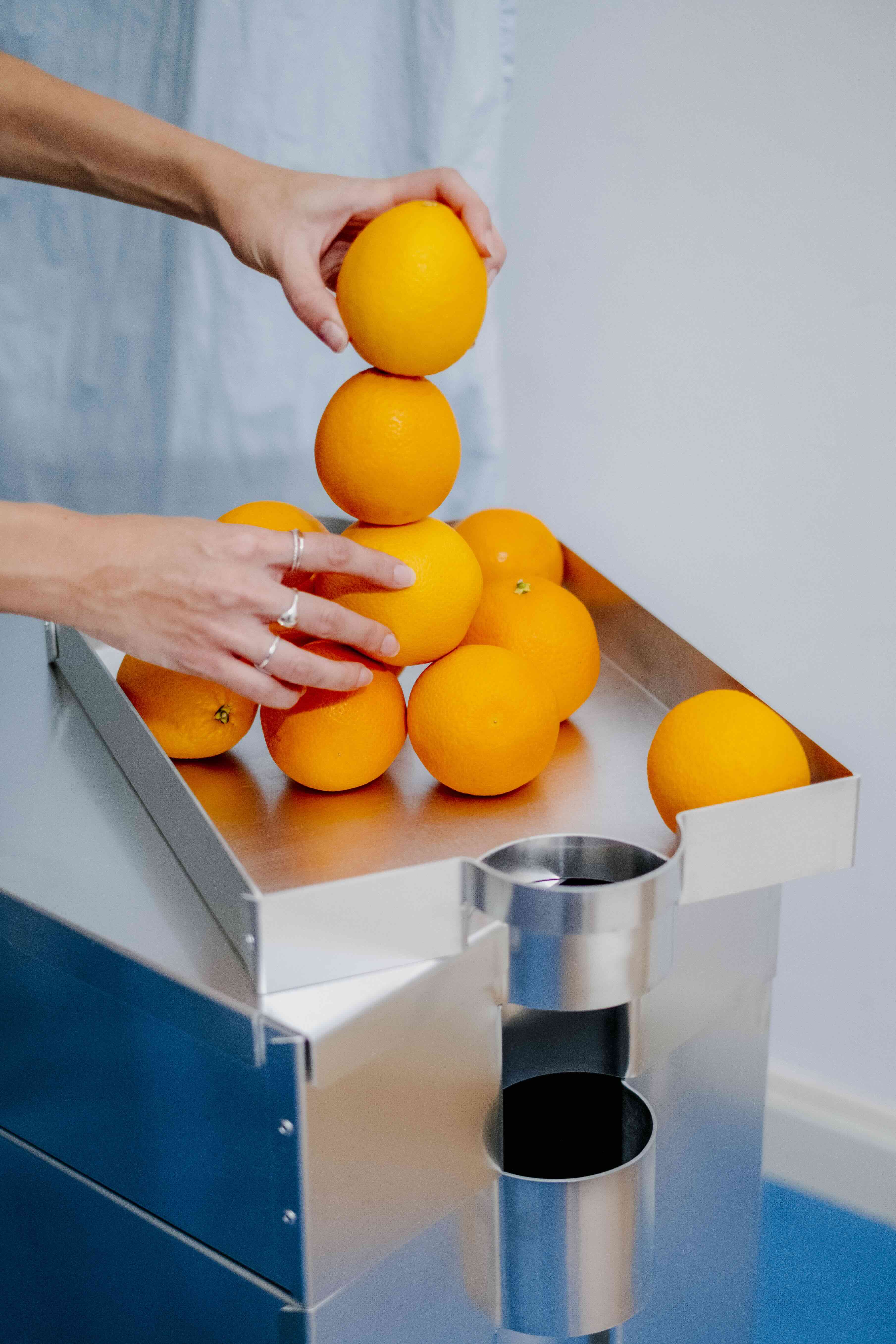 Hände stapeln Orangen auf einem minimalistischen Metallmöbel mit integrierten zylindrischen Öffnungen, modernes Produktdesign in Studio-Umgebung.