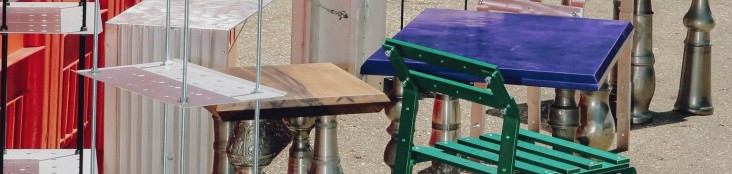 Verschiedene experimentelle Möbelstücke aus Metall und Holz in unterschiedlichen Farben, arrangiert im Außenraum vor industriellem Hintergrund.