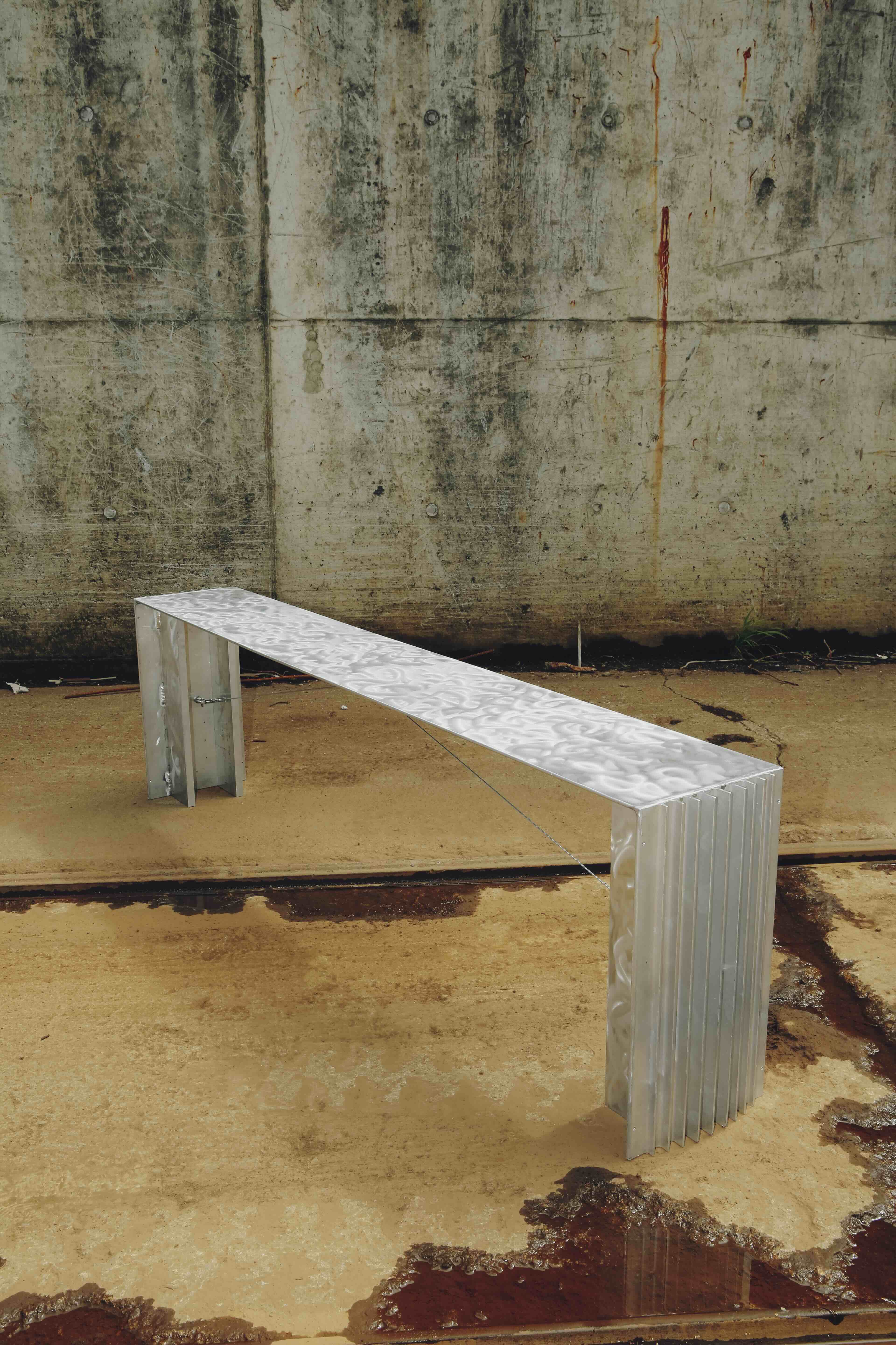 Minimalistische Sitzbank aus Metall vor einer rohen Betonwand, modernes Designobjekt in industrieller Umgebung.